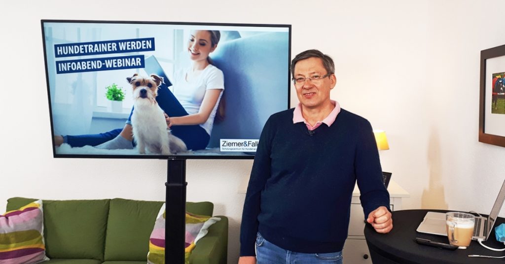 Ein lächelnder Mann mittleren Alters steht in einem Wohnzimmer neben einem großen Monitor, auf dem eine Frau mit einem kleinen Hund und der Schriftzug „Hundetrainer werden – Infoabend-Webinar“ zu sehen sind; auf einem Tisch daneben stehen ein Laptop und ein Glas Kaffee.