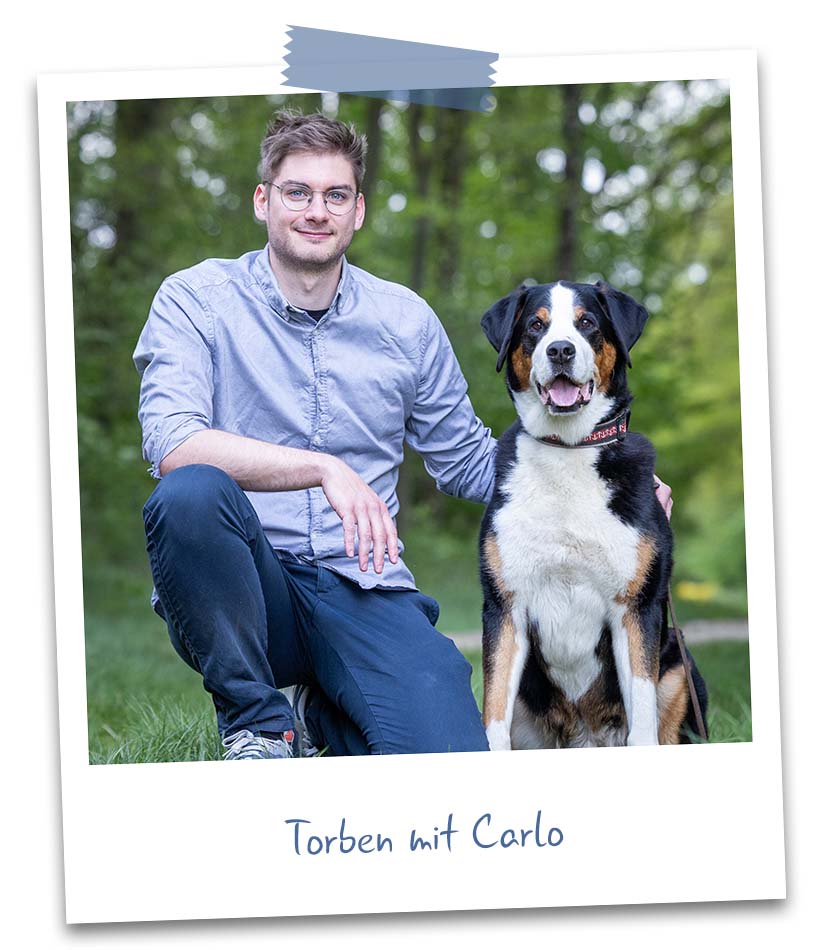 Ein junger Mann mit Brille kniet im Gras eines Parks neben einem großen dreifarbigen Hund, beide blicken entspannt in die Kamera.