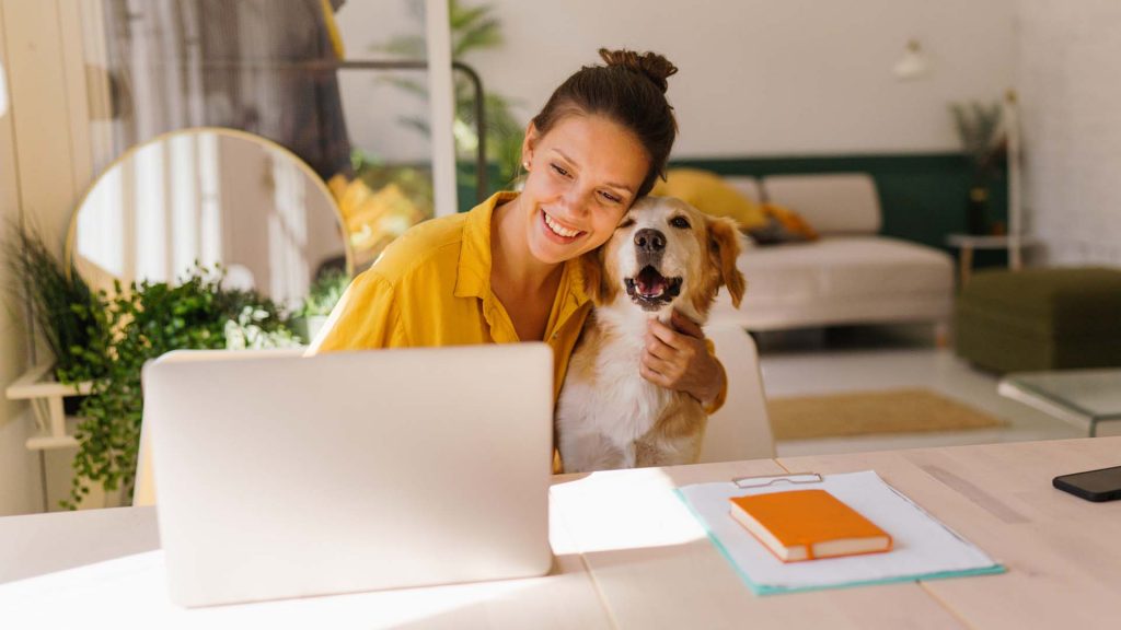 Ein junge Frau sitzt lächelnd zusammen mit einem Hund im Arm an einem Schreibtisch vor einem Laptop
