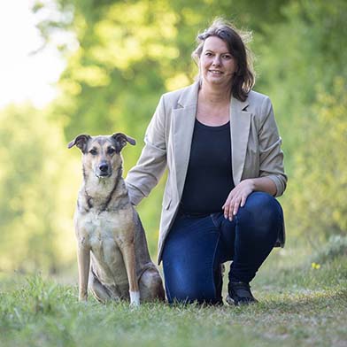 Frau mit schwarzem Top und beige-farbenem Blazer und Jeans hockt in grünem Freien und hält neben ihr einen sitzenden Hund im Arm.