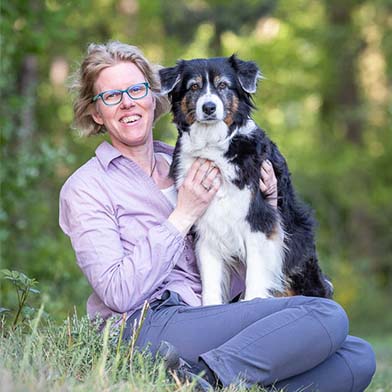 Frau in fliederfarbener Blus und blauen Jeans sitzt mit einem Australian Shepard auf dem Schoß in grünem Freien.