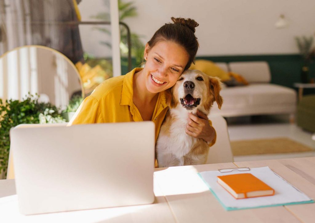 Ein junge Frau sitzt lächelnd zusammen mit einem Hund im Arm an einem Schreibtisch vor einem Laptop